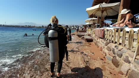 Blonde-Frau-Geht-Nach-Dem-Tauchen-Mit-Sonnenschirmen-Und-Bars-Entlang-Der-Meeresküste-Bei-Tageslicht-Von-Hinten-Gesehen-Am-ägyptischen-Strand-Spazieren