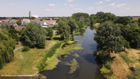 Flyover-about-stagnant-stream-running-through-village-in-Batya,-Hungary