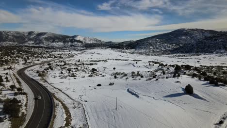 drone-flight-in-a-rural-area-with-a-significant-snowfall-we-see-a-road-and-some-vehicle-driving-with-a-blue-sky-with-clouds-and-a-background-of-mountains-that-cast-shadows-in-Avila-Spain