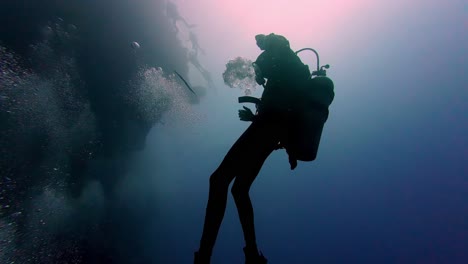 Ein-Taucher,-Dessen-Silhouette-Sich-Gegen-Das-Helle-Sonnenlicht-Abhebt,-Das-Durch-Die-Wasseroberfläche-Fällt,-Schwebt-In-Den-Tiefen-Des-Ozeans,-Umgeben-Von-Unzähligen-Kleinen-Blasen,-Die-Zur-Oberfläche-Aufsteigen.