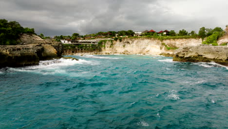 Drone-at-low-altitude-over-rough-sea-with-Blue-Lagoon-in-background,-Nusa-Ceningan,-Lembongan,-Bali-in-Indonesia