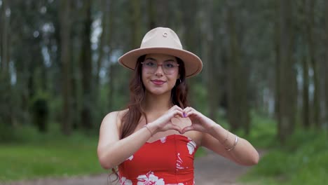 Ecuadorian-woman-in-a-vibrant-red-outfit,-adorned-with-glasses-and-a-cowboy-hat,-stands-amidst-the-forest-backdrop,-forming-a-heart-shape-with-her-hands-and-sharing-a-radiant-smile