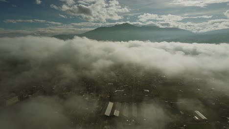 Aerial-view-of-the-town-of-Machachi-hidden-under-thick-clouds-and-fog,-the-dark-mountain-of-Pasochoa-volcano-in-the-distance