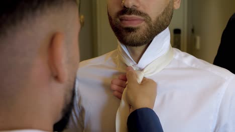 A-best-man-of-the-groom-puts-his-beige-tie-on-him,-over-his-white-shirt