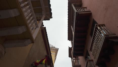 POV-Mit-Blick-Nach-Oben-Auf-Die-Balkone-Traditioneller-Kolonialhäuser-Mit-Blick-Nach-Unten-Auf-Die-Straße-Mit-Blick-Auf-Die-Kolumbianische-Flagge-An-Der-Wand