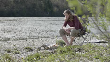 young-man-sitting-next-to-a-river-enjoying-the-bonfire