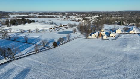 Aerial-view-of-a-snow-covered-landscape-with-patterned-fields,-trees,-a-pond,-and-clustered-buildings-with-a-smokestack