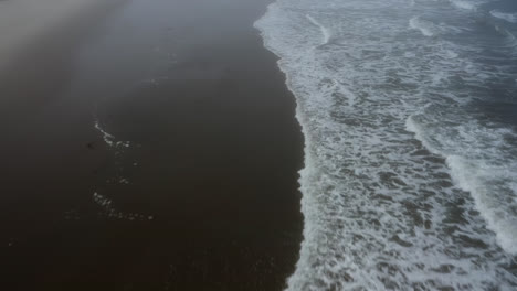 Foam-from-ocean-waves-rolling-onto-wet-sandy-beach,-Oregon-Coast,-low-aerial