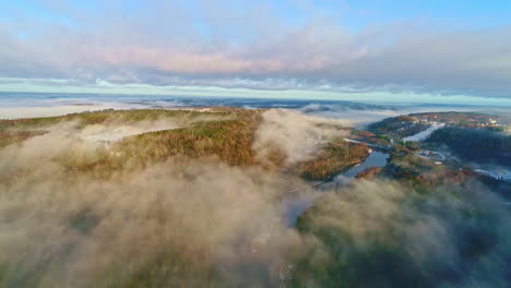 Drone-landscape-forested-hill-natural-area-with-a-river-crossing-between,-natural-valley,-flying-above-clouds-and-fog,-daily-skyline-background