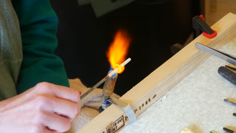 Artisan-heats-a-white-rod-with-a-blowtorch,-hands-in-focus,-against-a-wood-and-metal-workbench-backdrop