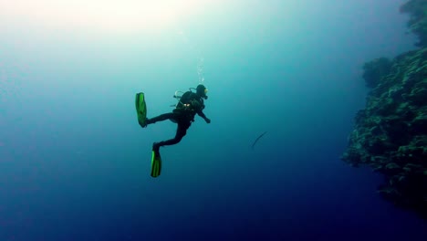 Taucher-Schwimmt-Inmitten-Der-Blauen-Unterwasser-Klippe-Im-Ozean-Und-Atmet-Blasen-In-Dahab,-Ägypten,-Mit-Panoramablick-Auf-Das-Sonnenlicht-über-Dem-Ozean