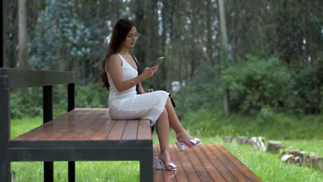 Latin-woman-in-a-white-dress-and-glasses,-seated-on-a-wooden-bench-in-the-forest,-as-she-enjoys-nature-while-taking-selfies-with-her-phone