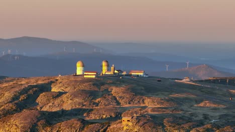 Vista-Aérea-Del-Amanecer-Con-Teleobjetivo-De-La-Antigua-Estación-De-Radar-Abandonada-Con-Turbinas-Eólicas-En-El-Fondo-En-La-Cima-Del-Parque-Natural-Serra-Da-Estrela,-Portugal