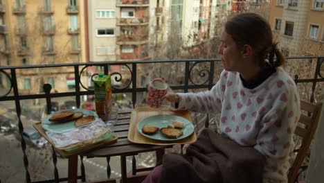 Gemütliche-Winterfrau,-Die-Im-Pyjama-Auf-Dem-Balkon-Sitzt,-Genießt-Frühstückstoast-Und-Eine-Tasse,-Während-Sie-Den-Straßenhintergrund-Beobachtet