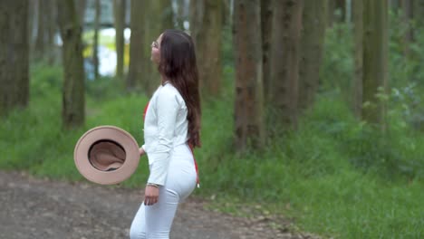 A-woman-in-white-attire-gracefully-walking-through-the-forest,-gently-removing-her-hat-amidst-towering-trees,-embodying-a-peaceful-escape-into-nature