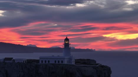 Luftdrohne-Umkreist-Leuchtturm,-Leuchtender-Lichtturm-Mit-Roten-Himmelsrichtungen,-Sonnenuntergang,-Skyline-Hintergrund,-Panorama-Reiseziel-Auf-Menorca,-Spanien
