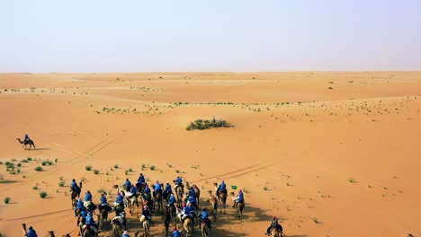 Vista-Aérea-Alrededor-De-Un-Safari-En-Camello-Por-El-Desierto,-árabes-Cabalgando-En-Un-Páramo-Sin-Fin,-Tarde-Soleada,-En-Arabia-Saudita---órbita,-Disparo-De-Drones