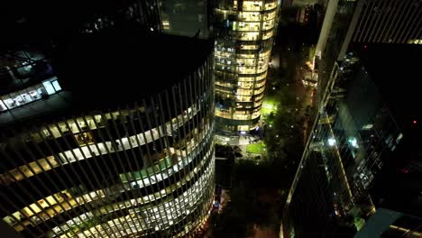 Aerial-drone-night-of-Santiago-de-Chile-business-offices-at-nueva-las-condes-neighborhood,-fly-around-lightened-architecture-with-windows-towards-city-streets