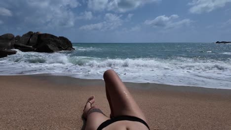 A-girl-resting-on-the-beach,-with-ocean-waves-gently-splashing-near-her-tanned-legs,-set-against-the-backdrop-of-the-paradise-island-of-Koh-Samui,-Thailand,-evoking-the-essence-of-travel-and-vacation