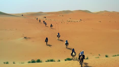 Aerial-view-over-people,-riding-camels-on-the-Arabian-desert,-during-golden-hour,-in-Saudi-Arabia---dolly,-drone-shot