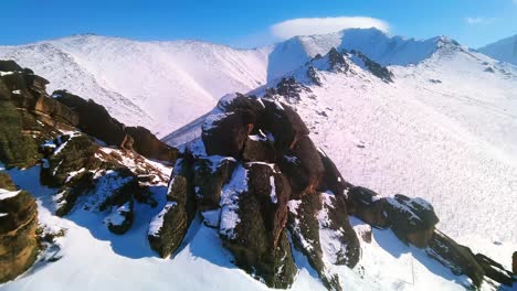 Beautiful-winter-rocks-against-the-backdrop-of-snow-capped-mountains-and-sunny-blue-sky-from-a-drone