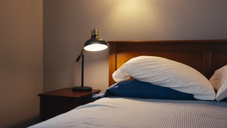 Eine-Nachttischlampe-Auf-Einem-Hölzernen-Nachttisch-Neben-Einem-Bett-Mit-Weißer-Bettwäsche-Und-Weißen-Und-Blauen-Kissen