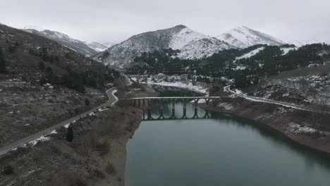 Picos-De-La-Cordillera-Congelada,-Puente-De-La-Zona-De-La-Carretera-Nevada-Que-Cruza-El-Río-Azul-Perspectiva-Aérea-De-Drones-En-El-Embalse