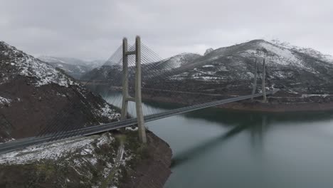 Bridge-road-crossing-mountain-range-above-river-winter-landscape-reservoir-of-Barrios-de-Luna-in-Leon-,-Spain-aerial-drone-orbit-view