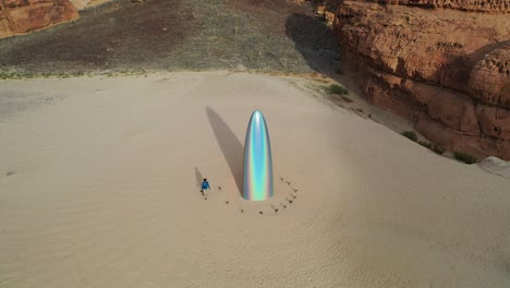 Aerial-view-of-a-man-walking-around-the-Gisela-Colon-parabolic-monolith,-at-the-Desert-x,-in-sunny-Al-ula,-Saudi-Arabia---descending,-tilt,-drone-shot