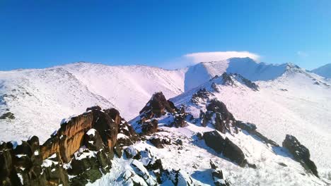 Beautiful-flyover-by-drone-near-winter-rocks-against-the-backdrop-of-snow-capped-mountains-and-sunny-blue-sky