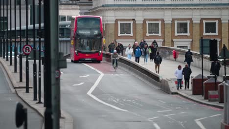 Ciclista-Seguido-Por-Un-Autobús-Londinense-En-El-Puente-De-Londres-Con-Multitudes-Caminando-Al-Fondo