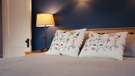 a-bed-with-white-pillows-with-artistic-designs-with-a-lamp-on-the-nightstand-all-in-front-of-a-blue-wall