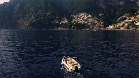 Disparo-De-Un-Dron-Siguiendo-Un-Barco-Que-Se-Movía-Hacia-El-Pueblo-De-Positano,-Hora-Dorada-En-Amalfi,-Italia