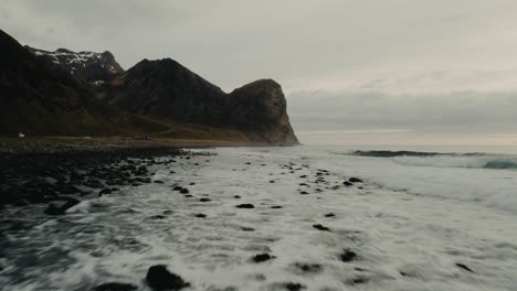 Waves-crashing-in-on-the-rocy-beach-surrounded-by-mountains-in-Lofoten-Islands-in-Norway-during-a-cloudy-day-and-grey-colors