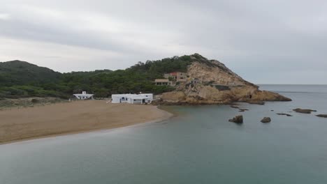 Aerial-drone-zooms-out-mediterranean-beach-location-in-menorca-Spain-on-a-cloudy-day