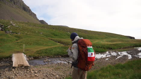 Man-walking-towards-a-small-bridge-and-a-stream,-summer-day,-in-Iceland---handheld,-slow-motion-view