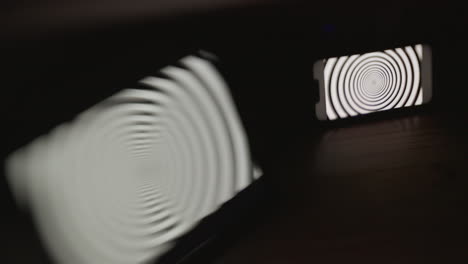 A-mobile-phone-screen-in-focus-and-tablet-out-of-focus-in-a-dark-room,-both-displaying-a-hypnotic-spiral-pattern