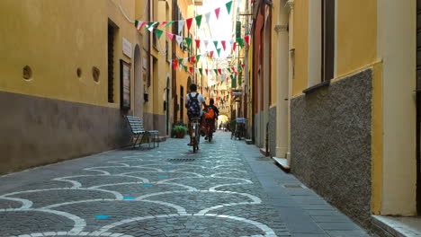 Drone-Siguiendo-A-Personas-En-Bicicleta-En-Un-Callejón-Estrecho-En-La-Soleada-Pietra-Ligure,-Italia