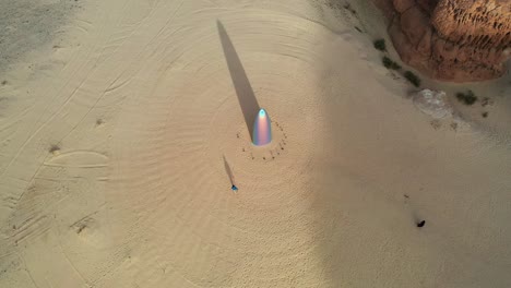 Aerial-view-of-a-man-walking-around-"the-future-is-now"-Gisela-Colon-parabolic-monolith,-at-the-Desert-x,-in-sunny-Al-ula,-Saudi-Arabia