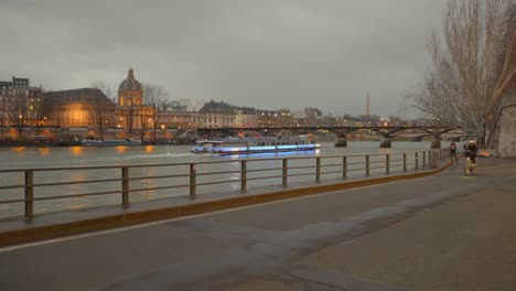 Toma-Panorámica-De-Una-Pista-Ciclista-Alrededor-Del-Río-Sena-Con-Un-Barco-Turístico-Y-El-Pont-Des-Arts-Al-Fondo.