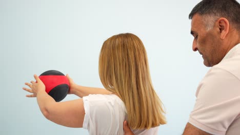 Physiotherapist-assists-blonde-woman-while-doing-shoulder-exercises-with-medicine-ball