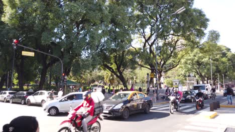 Motorcycles,-cars-drive-fast-by-Rivadavia-avenue-plaza-Pueyrredon-latin-traffic-in-bustling-city-of-Buenos-Aires-Argentine,-flores-neighborhood,-people-and-pedestrian