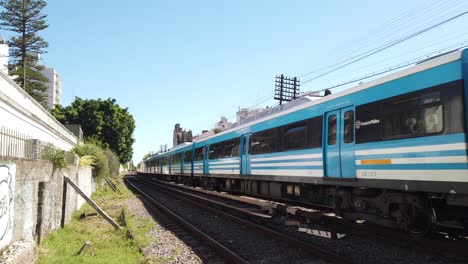 Blue-train-rides-Argentine-city-railway-Sarmiento,-wagons-pass-by-city-outskirt-in-buenos-aires,-public-transportation-service,-in-motion