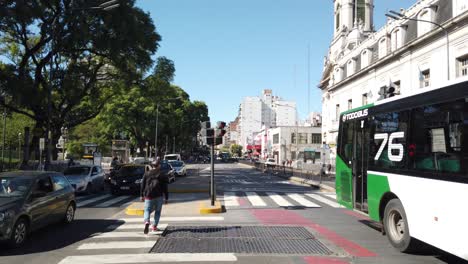 Elder-man-crosses-Rivadavia-avenue-bustling-city-of-Latin-America-traffic-park-urban-Landmark-of-buenos-aires-argentina,-buses,-cars-driving-along-Flores-Neighborhood,-plaza-Pueyrredon