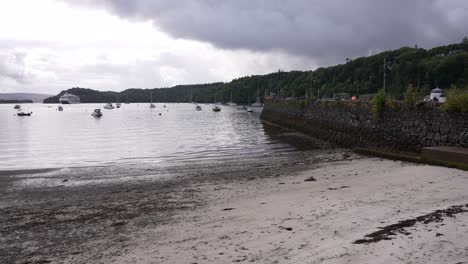 Tobermory,-Isle-of-Mull,-Scotland-UK,-Beach-Under-Coastal-Road,-Boats-and-Cruise-Ship-on-Cloudy-Day