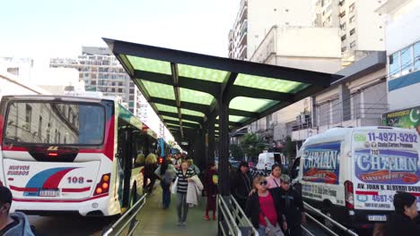 Bus-stop-people-commute-in-buenos-aires-argentina-south-American-bustling-city-autobus,-omnibus-platform,-station-at-flores-neighborhood,-argentine-pedestrians-in-commercial-neighborhood
