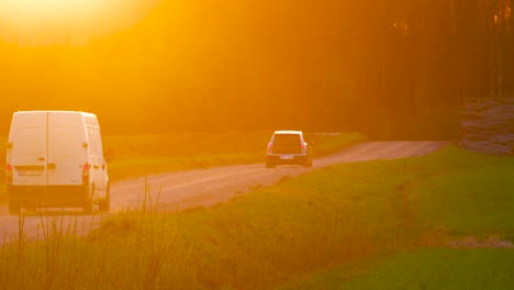 Cars-slowly-drive-on-scenic-countryside-gravel-road-during-bright-orange-sunset
