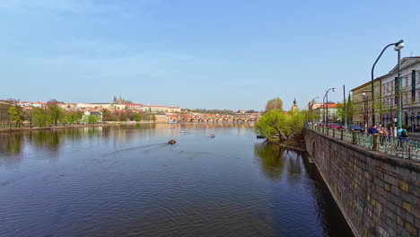 Shot-of-river-Vltava-flowing-through-the-city-of-Prague,-Czech-Republic-on-a-sunny-day-with-locals-passing-by-sidewalk