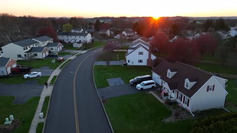 Sunset-starburst-highlights-spring-neighborhood-scene-with-homes-and-colorful-trees,-cars-on-street-at-intersection