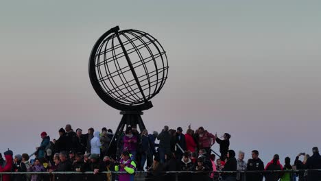 Drone-Shot-of-Tourist-Crowd-Around-Globe-Monument-Watching-Sunset-Above-North-Cape,-Norway-60fps